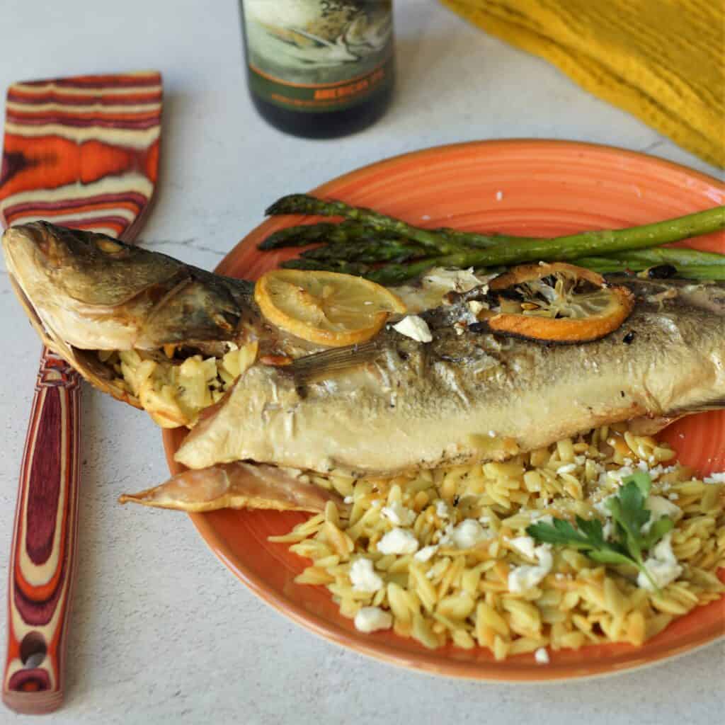 Stuffed European sea bass with orzo and feta on a plate with asparagus and a bottle of beer.