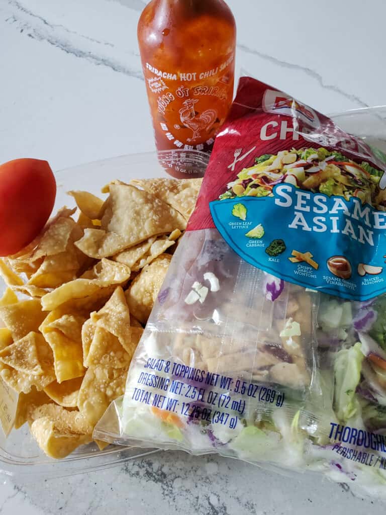 Asian salad mix, wonton, chips, a tomato, and sriracha sauce on a counter.