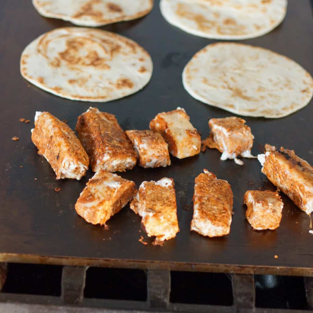 Pieces of seasoned mahi mahi and tortillas cooking on a flat top griddle.