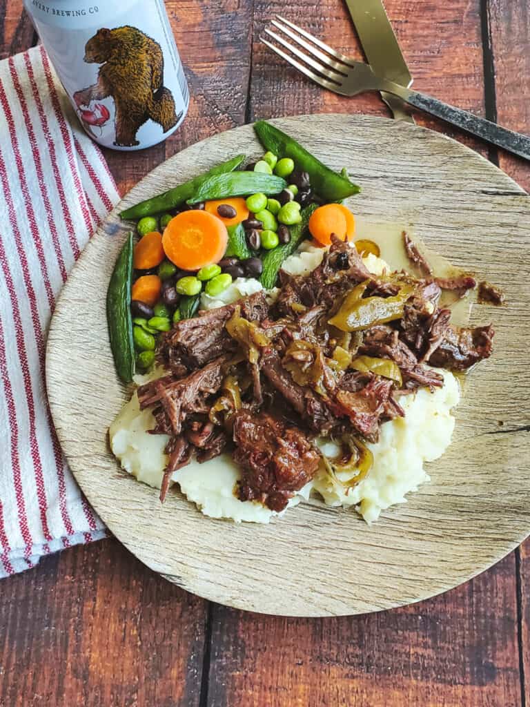 Mississippi Pot Roast served over mashed potatoes with vegetables on a wooden plate.