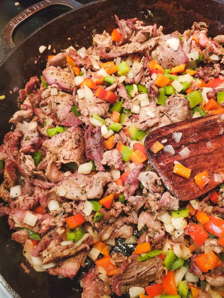 Thin sliced ribeye with peppers and onions for cheesesteaks being cooked in a cast iron pan.