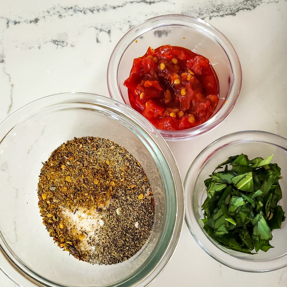 Diced Calabrian chilis, basil, and seasonings and herbs on a counter in small bowls.