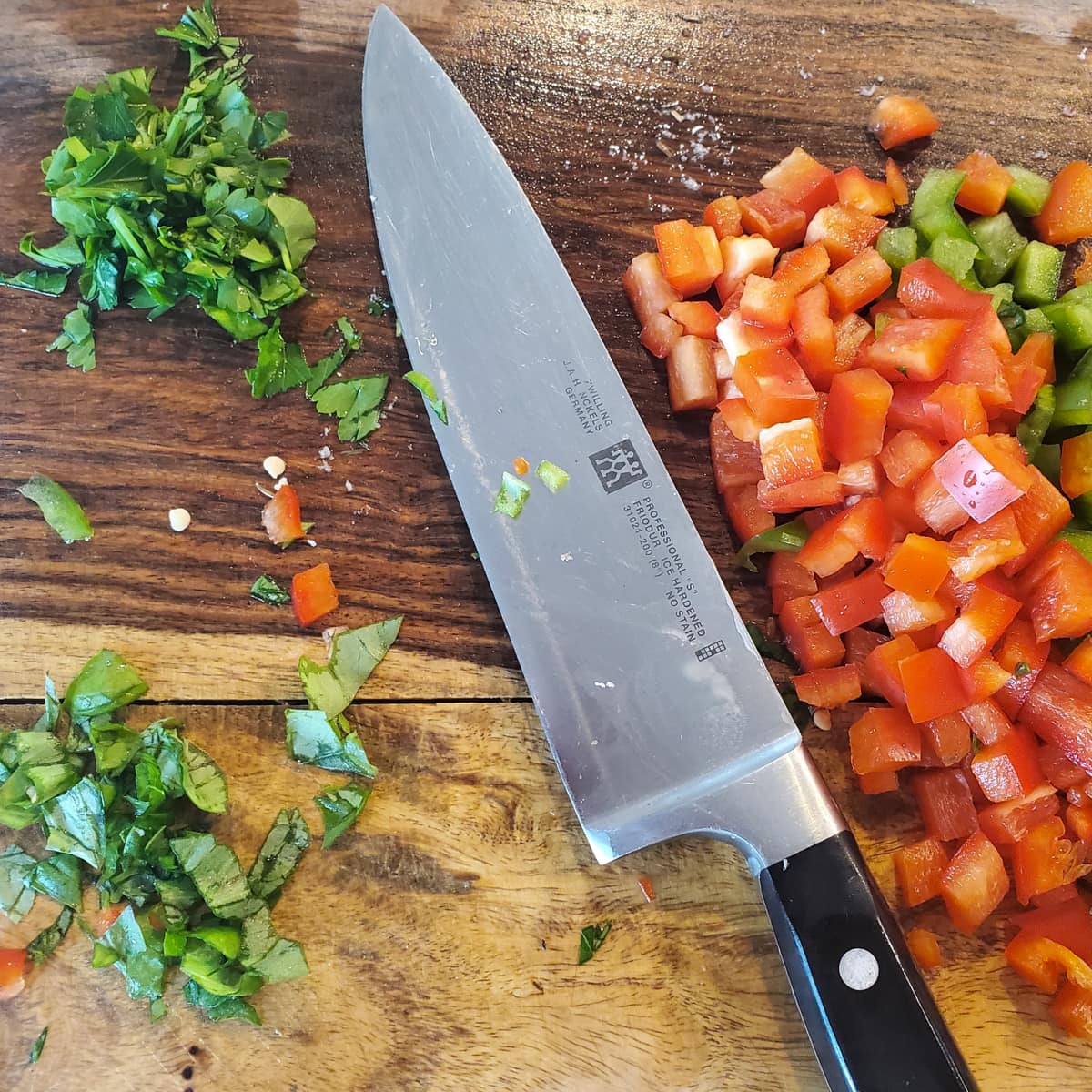 Diced bell peppers with chopped parsley and basil on a wood cutting board with a Chef's knife.