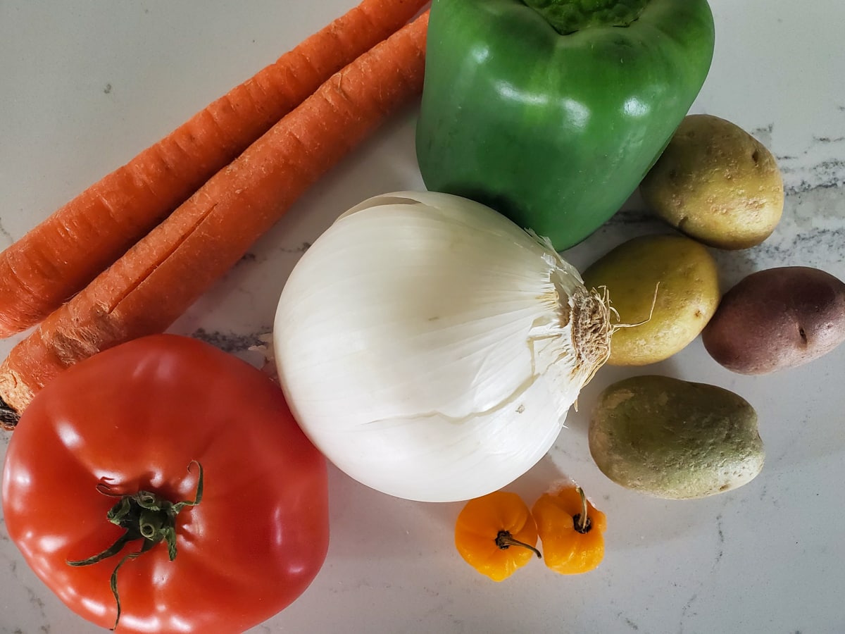 Bell pepper, carrot, tomato, potatoes and Scotch Bonnet peppers on a counter.