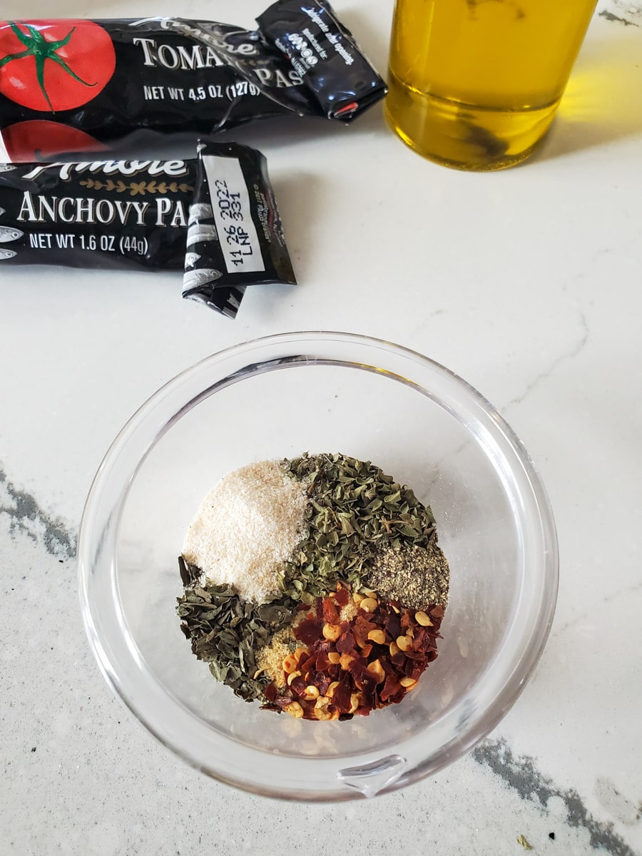 Spices and herbs for zesty pizza sauce and anchovy paste on a countertop.