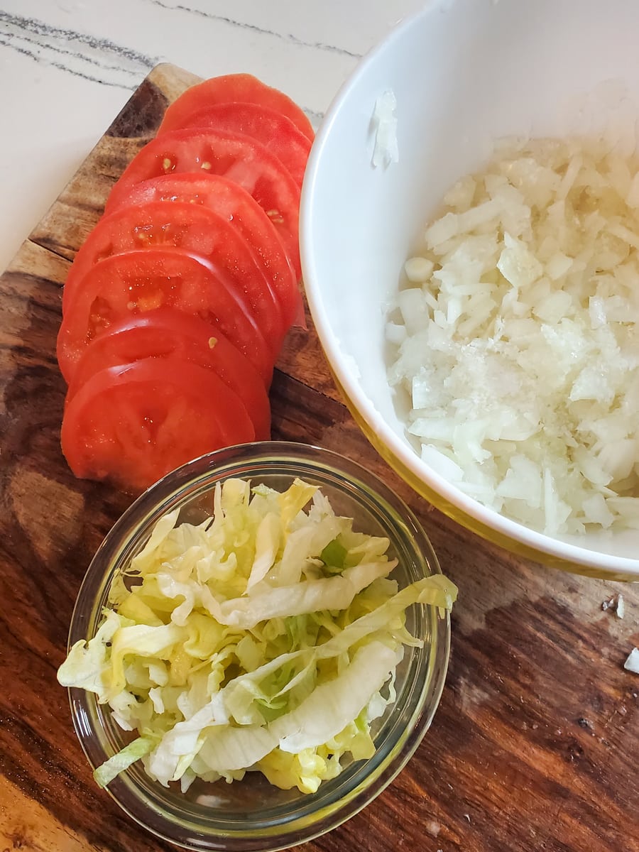 Wood cutting board with sliced tomatoes, shredded lettuce, and shredded onion.