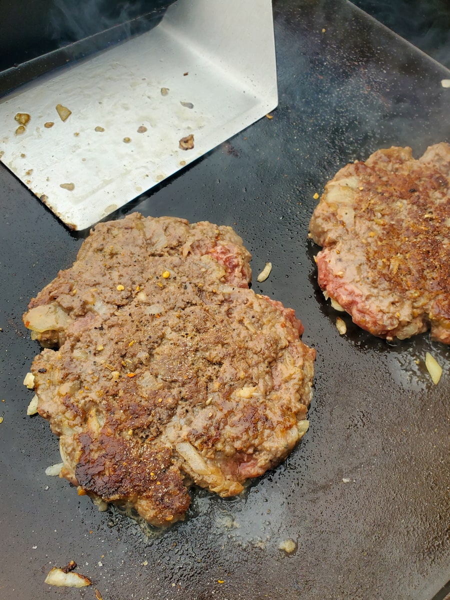 Seasoned burger patties cooking on a flat top griddle.