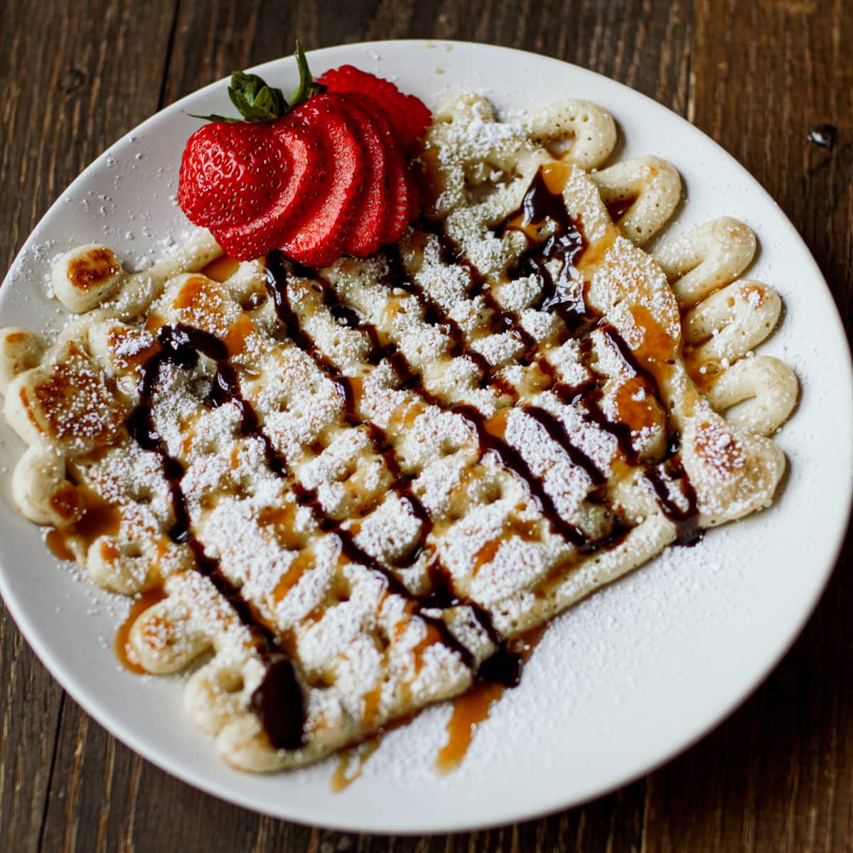 Homemade griddle funnel cakes with powdered sugar, chocolate syrup, and stawberries.
