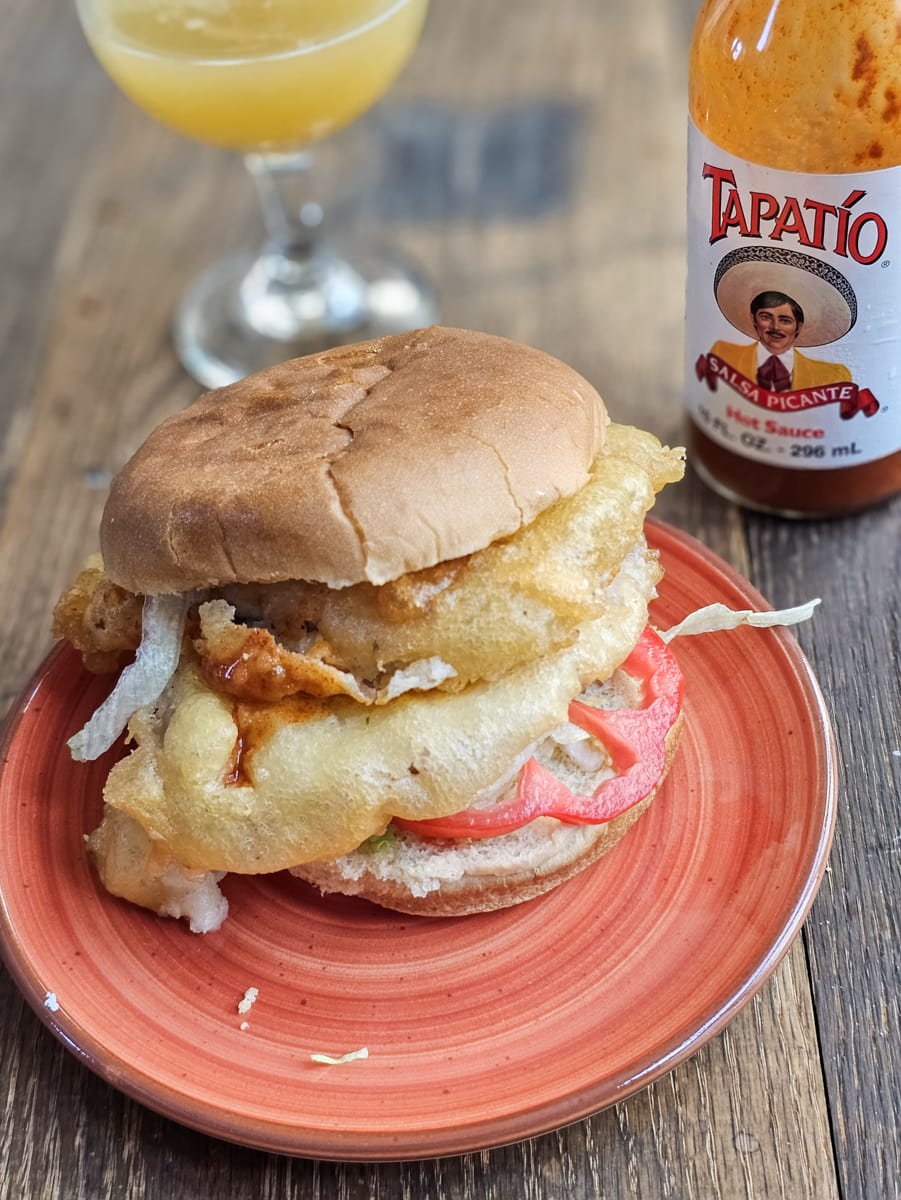 Fried tenderloin sandwich with tempura pork on an orange plate with beer and hot sauce.