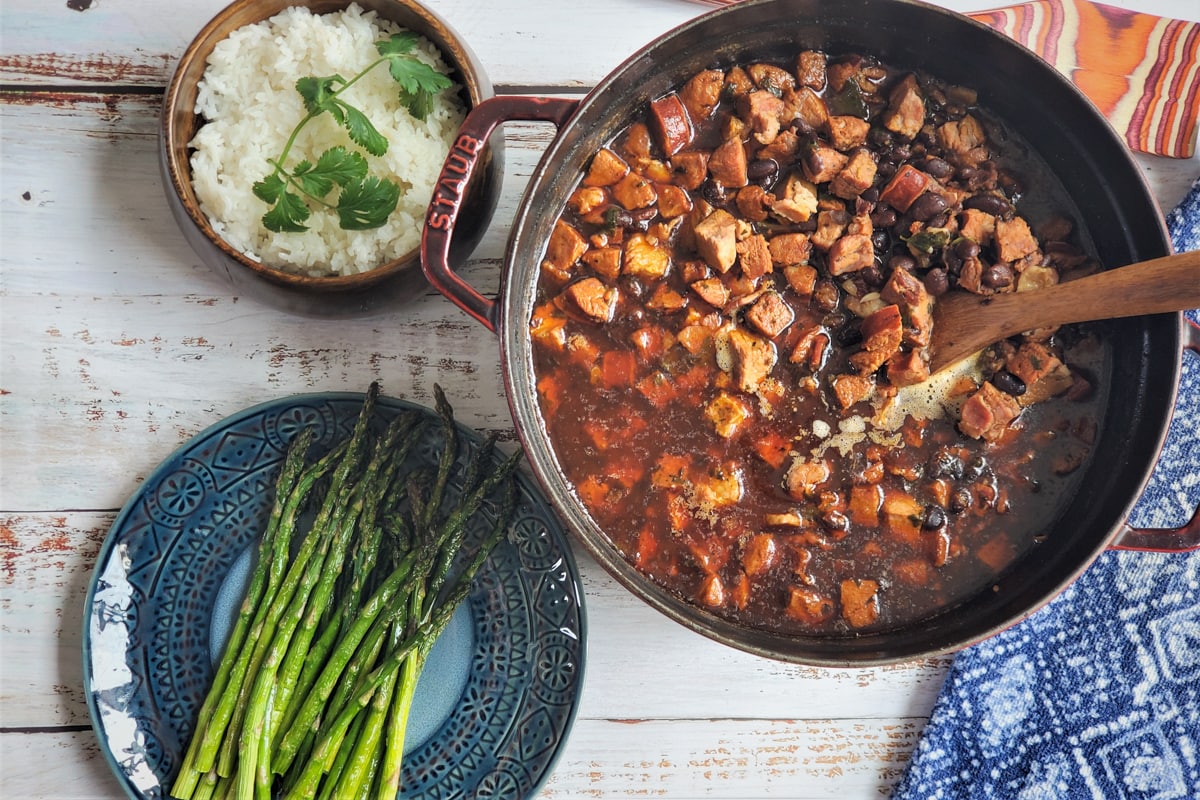 Staub Dutch Oven filled with Brazilian Feijoada next to a bowl of rice and plate of grilled asparagus.