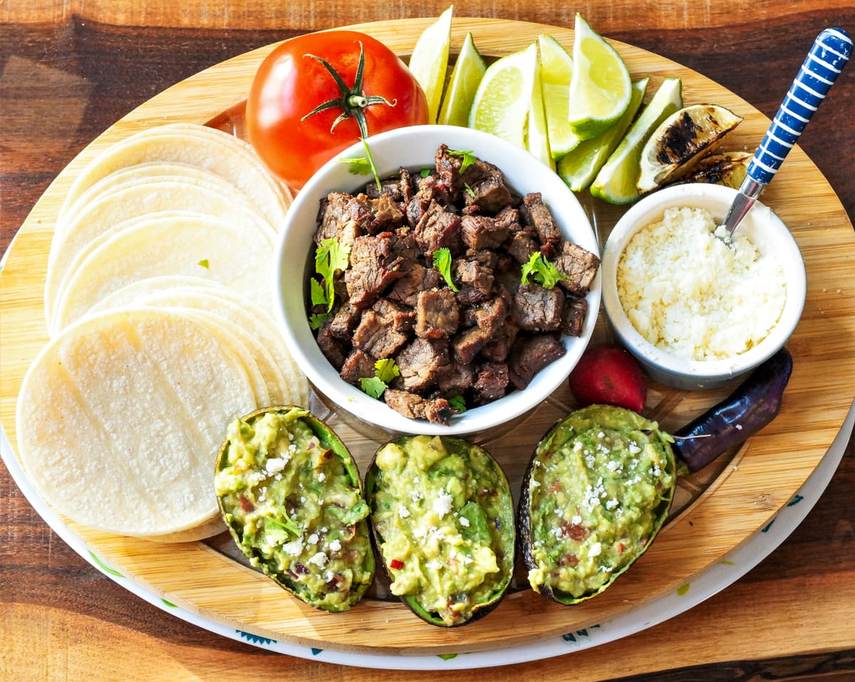 Carne Asada Tacos with tortillas, guacamole, lime wedges, and cheese.
