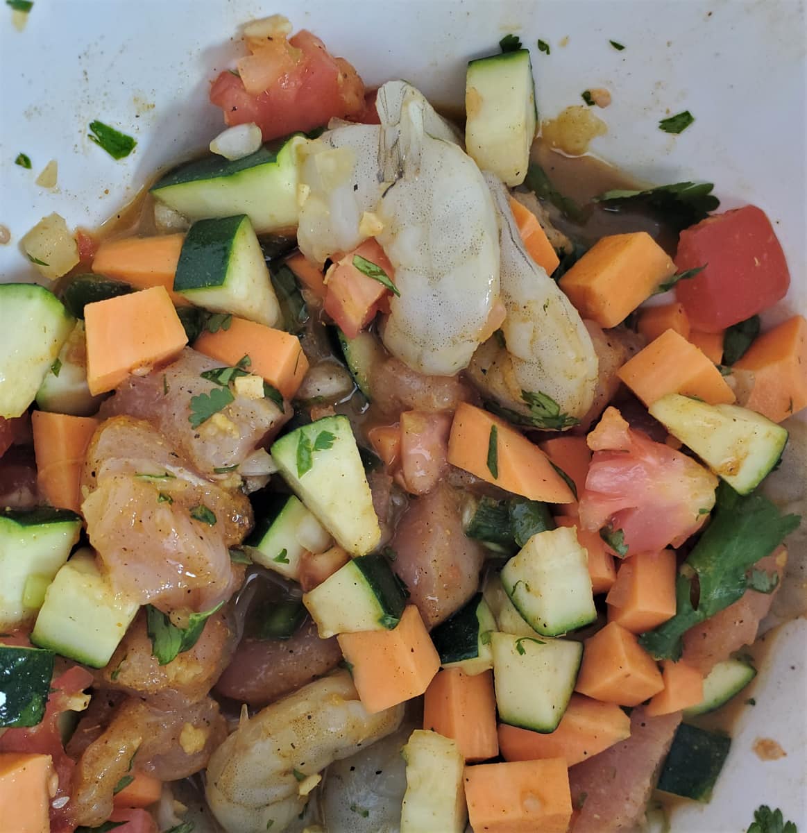 Close up of a bowl of diced onion, zucchini, garlic, tomato, and sweet potato with chicken and shrimp added.