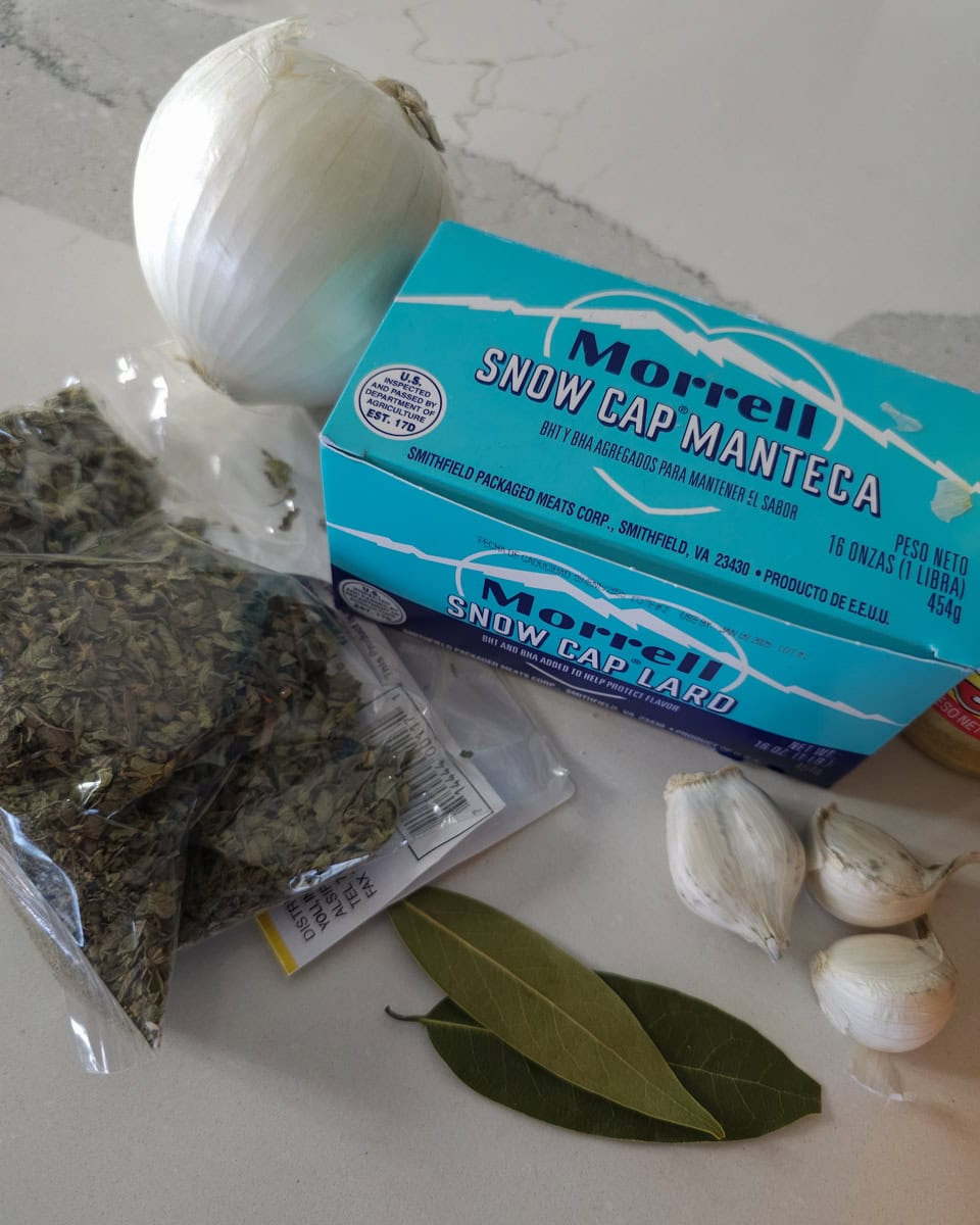 Lard, Mexican oregano, garlic, BAy leaves, and white onion on a countertop.