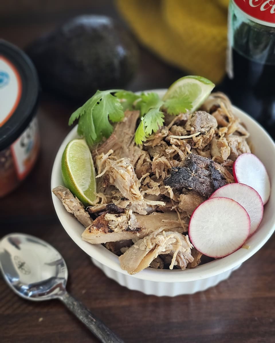 Bowl of homemade carnitas with lime wedges and radish slices.